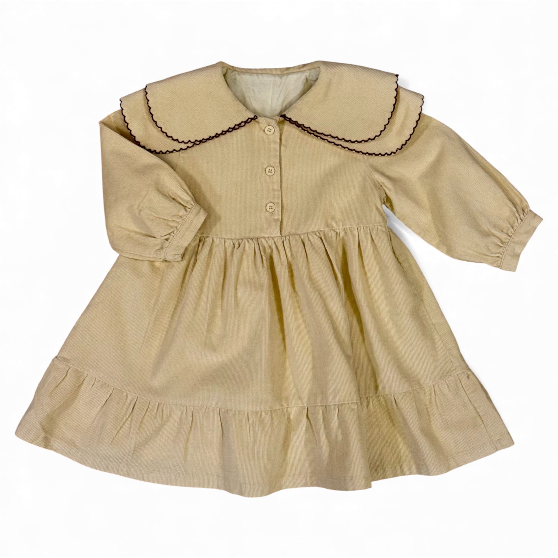 A beige needle-cord children’s dress with oversized Peter Pan bib collar, contrast scalloped trim, long balloon sleeves with gathered cuffs, empire line and full gathered skirt — front view.