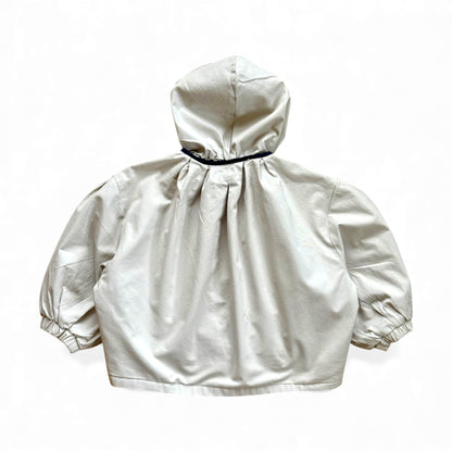 Reversible children’s jacket in plain cream organic cotton with floral lining, detachable hood, raglan sleeves and circular pockets — back view.
