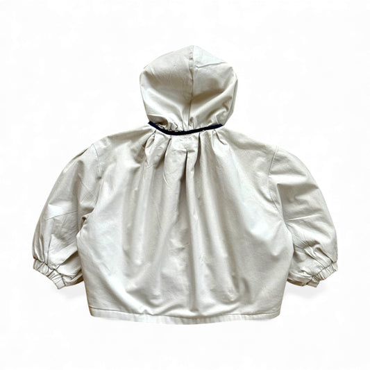 Reversible children’s jacket in plain cream organic cotton with floral lining, detachable hood, raglan sleeves and circular pockets — back view.