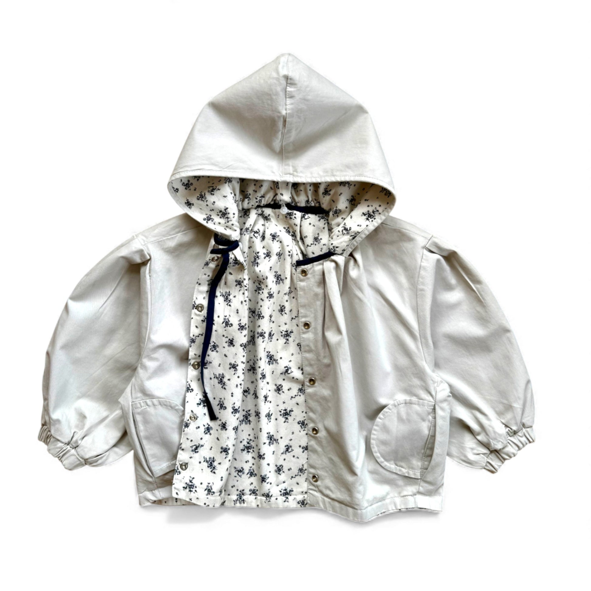 Reversible children’s jacket in plain cream organic cotton with floral lining, detachable hood, raglan sleeves and circular pockets — front view.