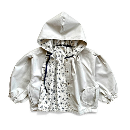 Reversible children’s jacket in plain cream organic cotton with floral lining, detachable hood, raglan sleeves and circular pockets — front view.