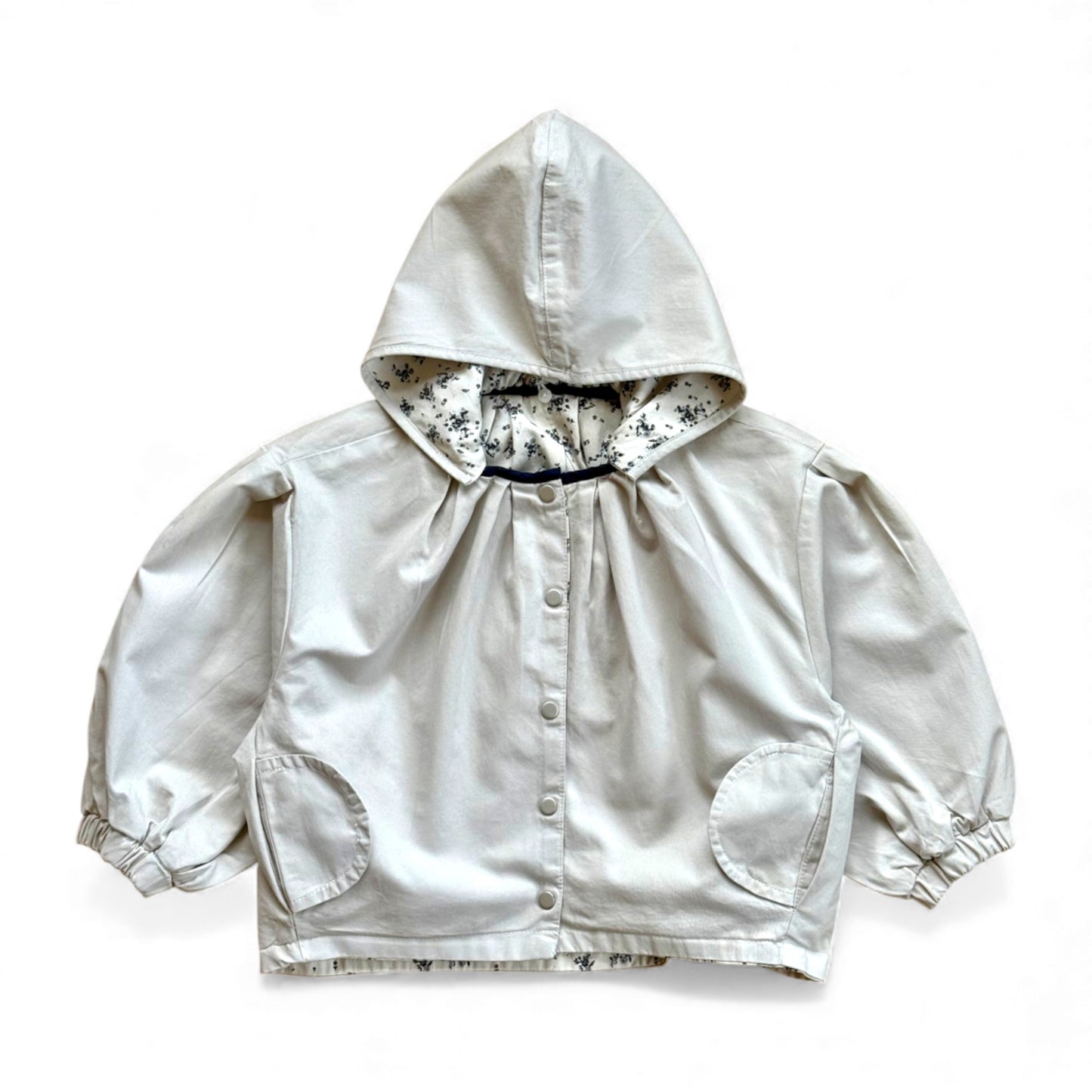Reversible children’s jacket in plain cream organic cotton with floral lining, detachable hood, raglan sleeves and circular pockets — front view.