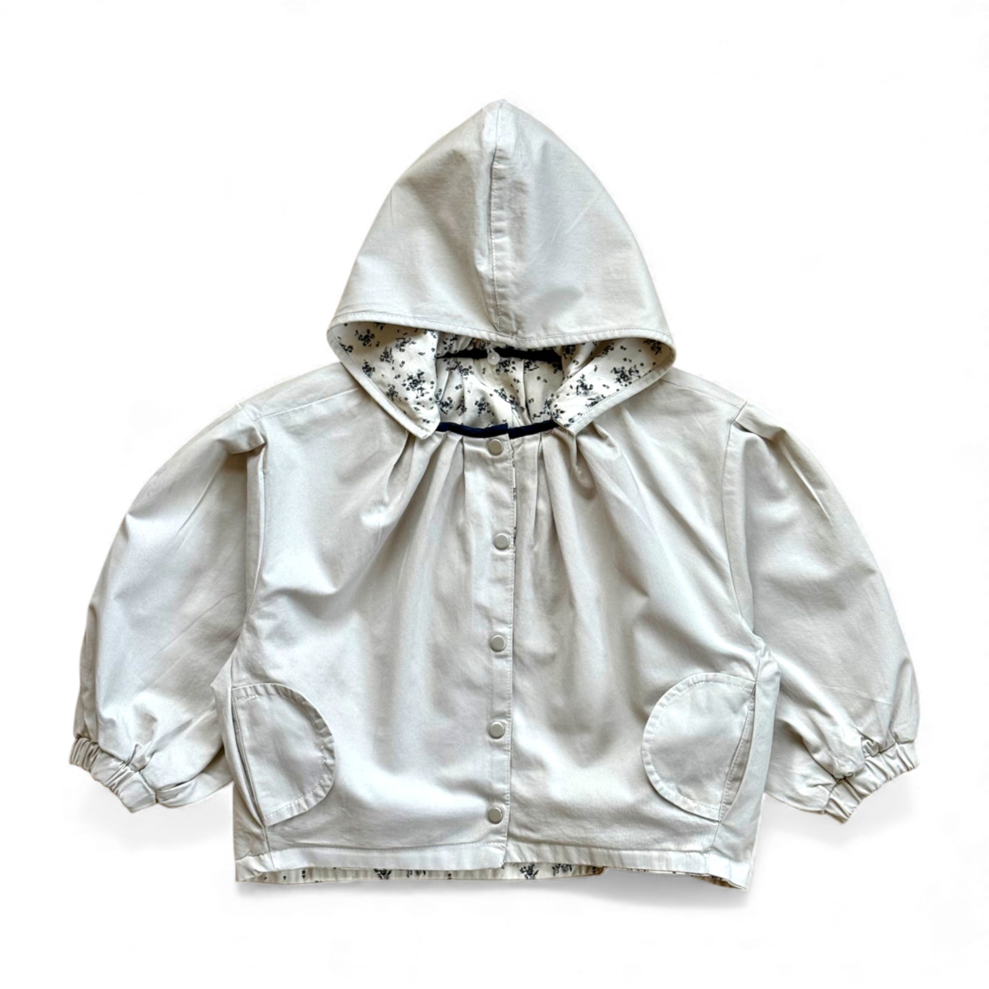 Reversible children’s jacket in plain cream organic cotton with floral lining, detachable hood, raglan sleeves and circular pockets — front view.
