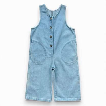 Light blue denim children’s romper with scoop neckline, circular front pockets and full front button closure
