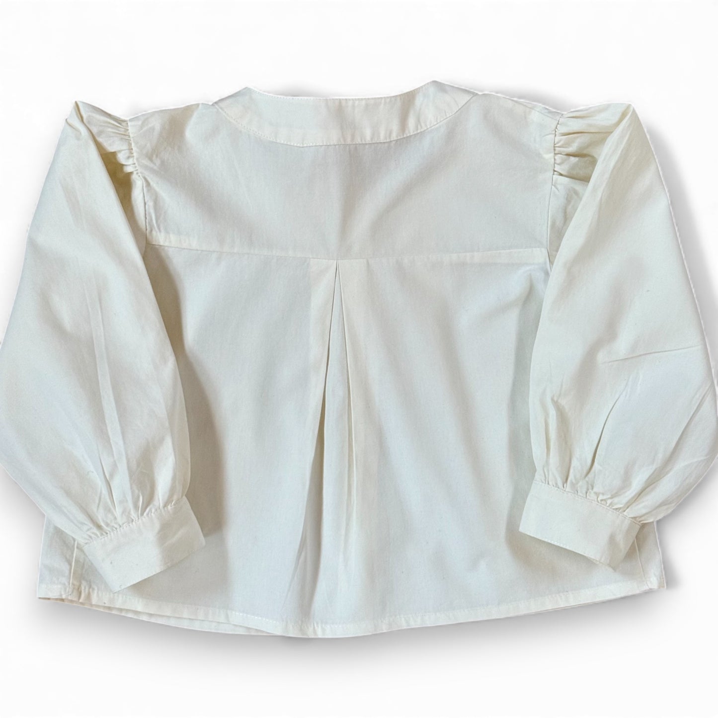Back view of children’s white A-line blouse with centre back pleat and gathered sleeves, laid flat.”
