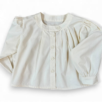 Children’s white A-line blouse with asymmetric band collar, off-centre pleats and drop shoulder, laid flat.